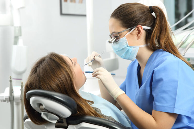 A dentist, wearing a blue surgical mask and gloves, is examining a patient's teeth with dental instruments.